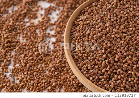 Organic buckwheat are scattered out of the wooden bowl on a light table, selective focus. Organic buckwheat are scattered out of the wooden bowl on a light table, selective focus. 112939727
