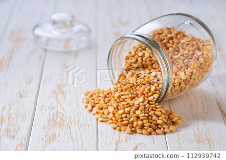 Raw dried peas spill out of a glass storage jar on a white wooden table. Raw dried peas spill out of a glass storage jar on a white wooden table. 112939742