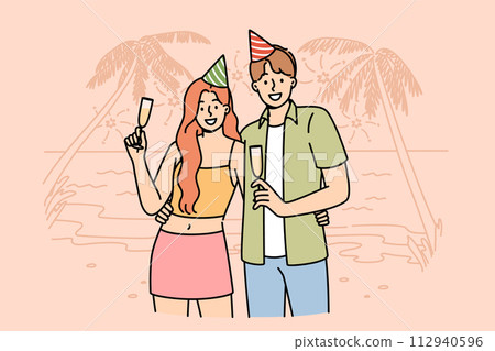 Honeymoon happy couple drinking champagne, standing on beach of sunny resort, wearing birthday caps 112940596