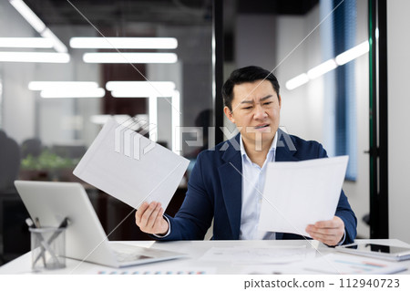 Punchy man holding documents while sitting at white desk in office environment. Handsome asian male doing work on background on glassy wall of business center. Concept of technology and people. 112940723