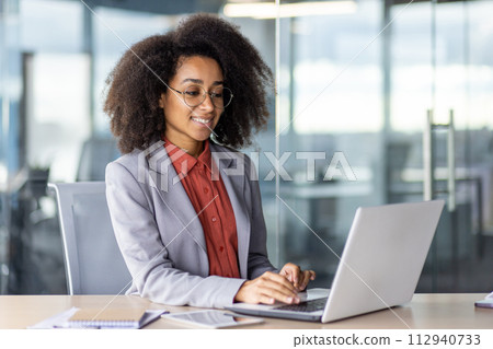 Side view of smiling hispanic woman with curly hair sitting at personal workplace and typing information on wireless laptop. Pretty lady using modern technologies for distance work in office interior. 112940733
