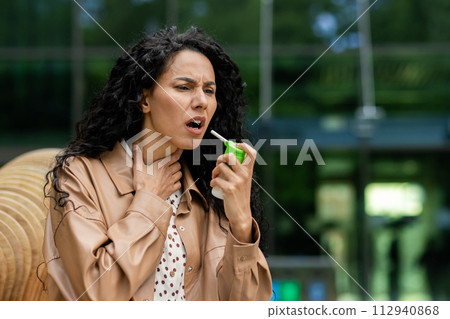 Upset female holding small green bottle and spraying throat spray into mouth to alleviating pain outdoors. Lady wearing light brown coat and shirt with brown spots on background of glassy building. Upset female holding small green bottle and spraying throat spray into mouth to alleviating pain outdoors. Lady wearing light brown coat and shirt with brown spots on background of glassy building. 112940868