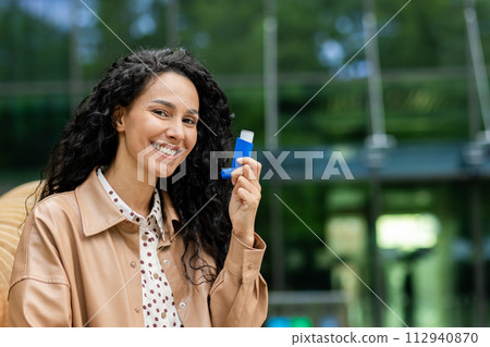 Smiling caucasian female holding blue inhaler close to face on background of green area and modern building. Beautiful woman making breathing easier while using nebulizer on fresh air. Smiling caucasian female holding blue inhaler close to face on background of green area and modern building. Beautiful woman making breathing easier while using nebulizer on fresh air. 112940870