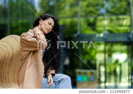 Side view of tired woman suffering from sharp pain in neck while having resting time on wooden bench near glassy building. Upset female with closed eyes massaging ache area outdoors. 112940877