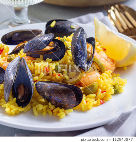Paella with mussels and shrimps, on white plate, with lemon and white wine, square, closeup 112943877