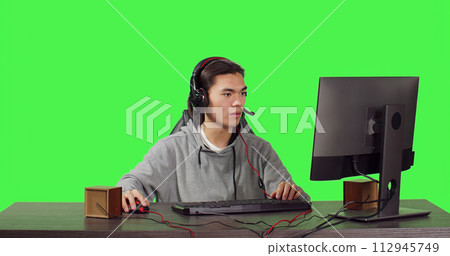 Young gamer sitting at computer desk and playing web based video games with people, using modern gadgets to have a good time. Asian person competing in rpg contest over greenscreen. 112945749