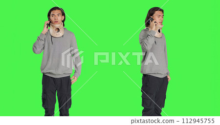 Young adult with neck collar answers call on smartphone, talking to people about his bone fracture accident. Hurt person wearing cervical foam brace discussing on telephone line, greenscreen. 112945755