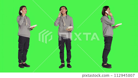 Asian guy talking on landline phone, answering call on telephone with cord while he stands against full body greenscreen. Young person using retro office phone for remote communication. Asian guy talking on landline phone, answering call on telephone with cord while he stands against full body greenscreen. Young person using retro office phone for remote communication. 112945807