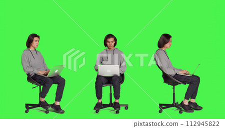 Young adult browsing the web on laptop in full body greenscreen studio, using mobile computer to navigate through webpages online. Person sitting on chair and working on wireless pc, trendy guy. 112945822
