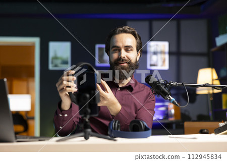 Cheerful content creator endorsing headphones for online streaming platforms viewers, filming with phone. Man promoting music listening device, doing influencer marketing using smartphone on tripod Cheerful content creator endorsing headphones for online streaming platforms viewers, filming with phone. Man promoting music listening device, doing influencer marketing using smartphone on tripod 112945834