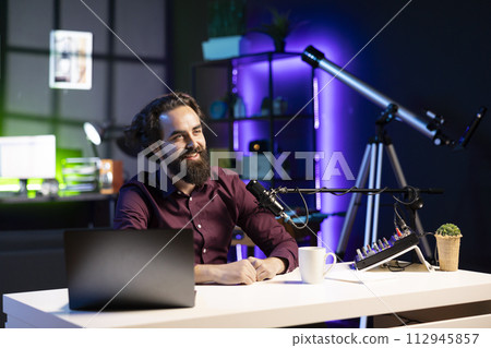 Happy internet star using quality mic in studio to discussing with viewers. Smiling man sitting at table in front of laptop using high tech recording gear to record social media video Happy internet star using quality mic in studio to discussing with viewers. Smiling man sitting at table in front of laptop using high tech recording gear to record social media video 112945857