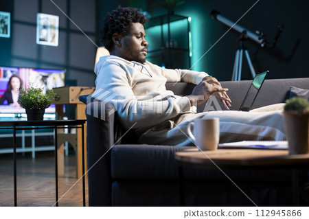 Self employed man inputting data on laptop while working from home, looking at screen. BIPOC person lying on couch, remotely performing tasks in nice stylish warm apartment living room 112945866