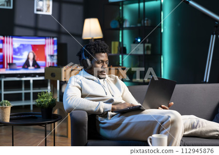 African american man feeling bored, scrolling on internet using laptop while listening to music, relaxing during weekend. Person wearing headphones, surfing the net at home 112945876