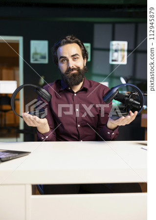 Content creator in neon lit home studio filming wireless headphones review for online streaming platforms. Media star hosts internet show, comparing music listening devices from different price ranges 112945879