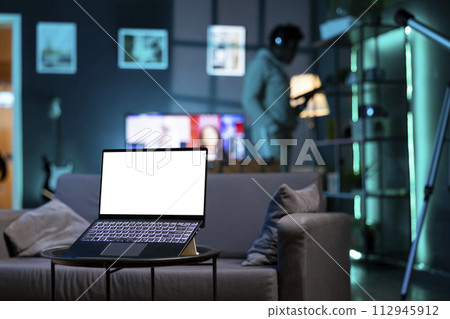 Focus on isolated screen laptop on coffee table in apartment with man in blurry background cleaning dust on shelves. Close up shot of mockup notebook and person in the back with tablet in hand Focus on isolated screen laptop on coffee table in apartment with man in blurry background cleaning dust on shelves. Close up shot of mockup notebook and person in the back with tablet in hand 112945912