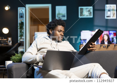 Remote employee concentrating on finishing up important job tasks, reading files info in apartment personal office. Focused teleworker crosschecking documents between laptop and paperwork 112945927