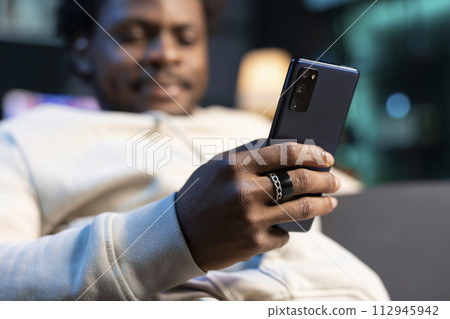 Close up of cellphone held by person chatting with online friends, enjoying himself at home. Focus on mobile phone used by BIPOC man in blurry background to scroll on social media 112945942