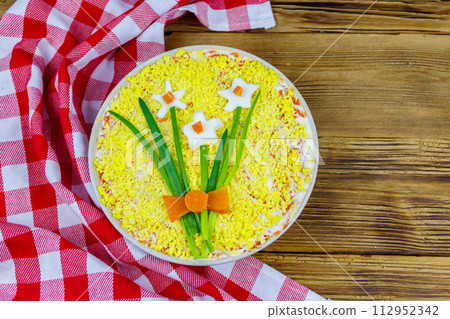 Traditional russian layered salad Mimosa with spring decoration Daffodil on wooden table. Top view. Decoration is made of egg, green onion and carrot 112952342