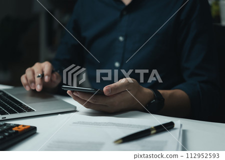 Businessman hand holding mobile smartphone on a table with a laptop at office.... 112952593