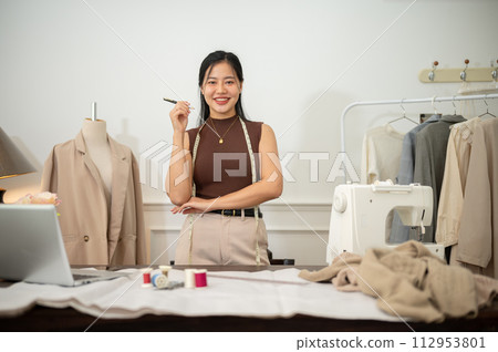An attractive Asian female fashion designer or tailor is in her studio, smiling at the camera. An attractive Asian female fashion designer or tailor is in her studio, smiling at the camera. 112953801