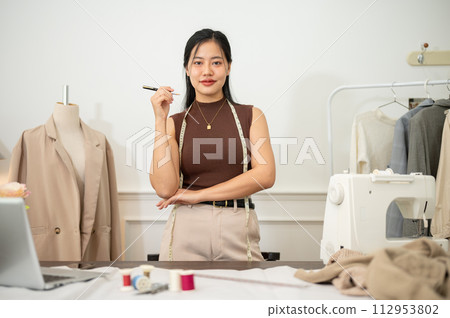 An attractive Asian female fashion designer or tailor is in her studio, smiling at the camera. An attractive Asian female fashion designer or tailor is in her studio, smiling at the camera. 112953802