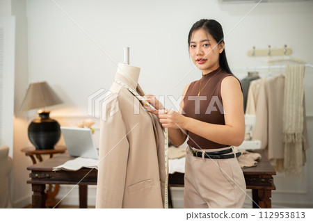 A professional Asian female tailor is working in her studio, measuring sizes with a measuring tape. A professional Asian female tailor is working in her studio, measuring sizes with a measuring tape. 112953813