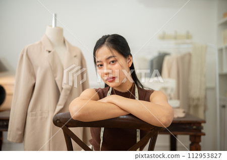 A confident Asian female fashion designer or tailor is sitting on a chair in her studio. 112953827