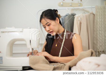 A professional Asian female tailor or fashion designer is sewing a dress with a sewing machine. A professional Asian female tailor or fashion designer is sewing a dress with a sewing machine. 112953836