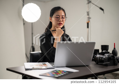 A confident Asian female photographer is working on her laptop, working in a photoshoot studio. 112954105
