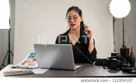 A confident Asian female photographer is working on her laptop, working in a photoshoot studio. A confident Asian female photographer is working on her laptop, working in a photoshoot studio. 112954107