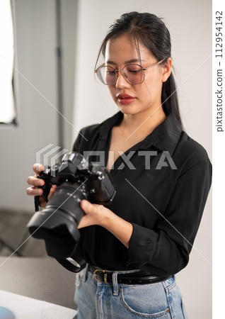 A confident, experienced Asian female photographer is adjusting her DSLR camera, preparing photoshoot equipment, and working in the studio. 112954142