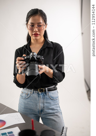 A confident, experienced Asian female photographer is adjusting her DSLR camera, preparing photoshoot equipment, and working in the studio. A confident, experienced Asian female photographer is adjusting her DSLR camera, preparing photoshoot equipment, and working in the studio. 112954144