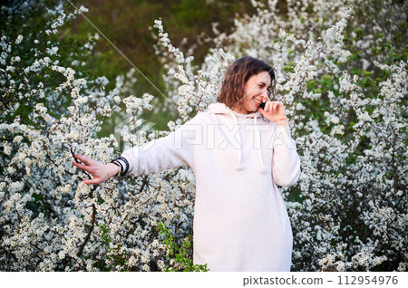 Woman allergic suffering from seasonal allergy at spring in blossoming garden at springtime. Young woman sneezing, closing nose by hand in front of blooming tree. Spring allergy concept 112954976