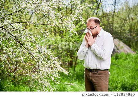 Man allergic suffering from seasonal allergy at spring in blossoming garden at springtime. Young man sneezing and blowing nose with nasal handkerchief in front of blooming tree. Spring allergy concept Man allergic suffering from seasonal allergy at spring in blossoming garden at springtime. Young man sneezing and blowing nose with nasal handkerchief in front of blooming tree. Spring allergy concept 112954980