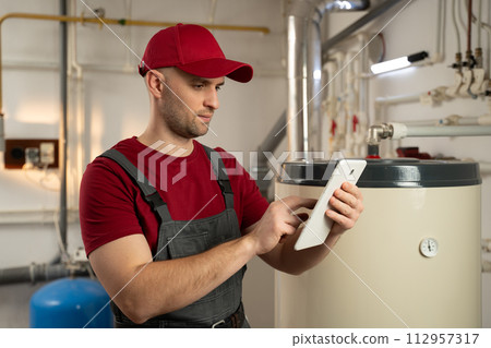 Professional Plumber Inspects Water Heater in Residential Basement During Daytime 112957317
