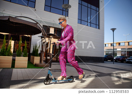 Stylishly dressed mature woman in a bright pink pantsuit of trousers and a jacket rides around the city on a scooter, an active, healthy and eco friendly lifestyle Stylishly dressed mature woman in a bright pink pantsuit of trousers and a jacket rides around the city on a scooter, an active, healthy and eco friendly lifestyle 112958583