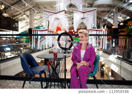 Beautiful mature blonde woman with a short haircut, an influencer, records a video blog while sitting at a table in a restaurant. Middle aged female blogger Beautiful mature blonde woman with a short haircut, an influencer, records a video blog while sitting at a table in a restaurant. Middle aged female blogger 112958586