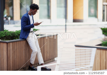Stylish African American man in smart casual clothes, with mobile phone against backdrop of city. Fashionable guy with smartphone outdoors Stylish African American man in smart casual clothes, with mobile phone against backdrop of city. Fashionable guy with smartphone outdoors 112958587