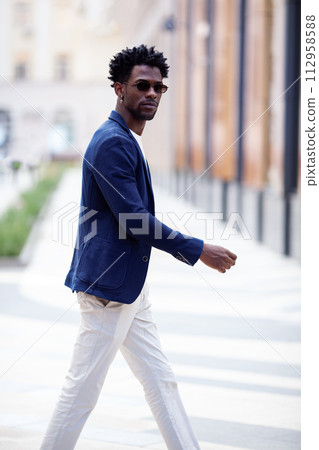 Handsome fashion African American man walks down the street in stylish attire white pants, blue blazer. Fashionable street style trend for men 112958588