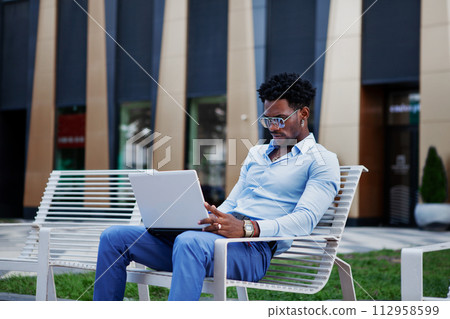 Young african american business man using laptop while sitting on bench in city street. Lifestyle of professional male working in urban environment Young african american business man using laptop while sitting on bench in city street. Lifestyle of professional male working in urban environment 112958599