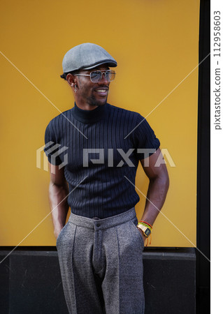 Handsome stylish African American man stands on background of yellow wall outdoors in fashionable attire, wearing cap, sunglasses, black t-shirt and trousers with arrows, modern male fashion model Handsome stylish African American man stands on background of yellow wall outdoors in fashionable attire, wearing cap, sunglasses, black t-shirt and trousers with arrows, modern male fashion model 112958603