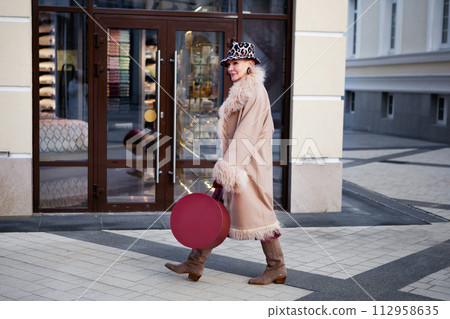 Fashionable mature woman walking in the city, dressed in a long beige coat, Cossack boots, hat with animal print and original handbag box. Street style trend for mature women 112958635