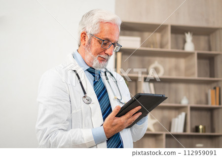 Mature senior male doctor in medical uniform using digital tablet in clinic. Therapist professional healthcare expert searching information online in hospital room. Medicine healthcare medical checkup 112959091