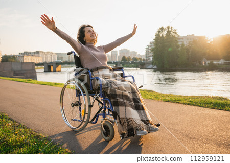 Happy senior woman in wheelchair raised hands enjoying freedom walking on road in city park outdoor. Old happy woman in chair for people with disability has a hope. Disability disease rehabilitation Happy senior woman in wheelchair raised hands enjoying freedom walking on road in city park outdoor. Old happy woman in chair for people with disability has a hope. Disability disease rehabilitation 112959121