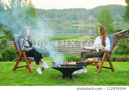 Two women in the backyard, sitting in chairs and chatting near the barbecue with vegetables Two women in the backyard, sitting in chairs and chatting near the barbecue with vegetables 112959504