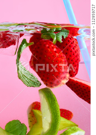 Sliced strawberries, lime and green leaves floating in water with light blue straw against pink background. Texture of cooling sweet summer's drink. Sliced strawberries, lime and green leaves floating in water with light blue straw against pink background. Texture of cooling sweet summer's drink. 112959717