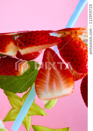 Sliced strawberries, carambola and green leaves floating in water with light blue straw against pink background. Sliced strawberries, carambola and green leaves floating in water with light blue straw against pink background. 112959728