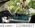 Happy Man Loading Potted Flowers into Car Trunk in Summer 112960827
