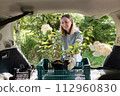 Happy Woman Loading Potted Flowers into Car Trunk in Summer 112960830