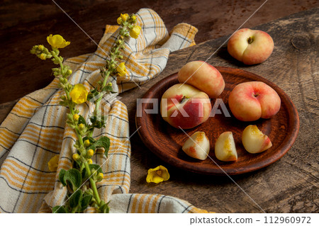 Clay plate with cut and whole saturn peaches or flat peaches on wooden background with yellow kitchen towel.. Clay plate with cut and whole saturn peaches or flat peaches on wooden background with yellow kitchen towel.. 112960972